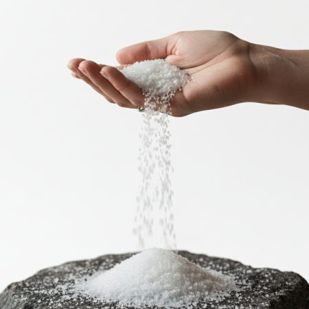 Mano femenina dejando caer cristales de sal blanca sobre una roca oscura, simbolizando pureza y el origen de la haloterapia.